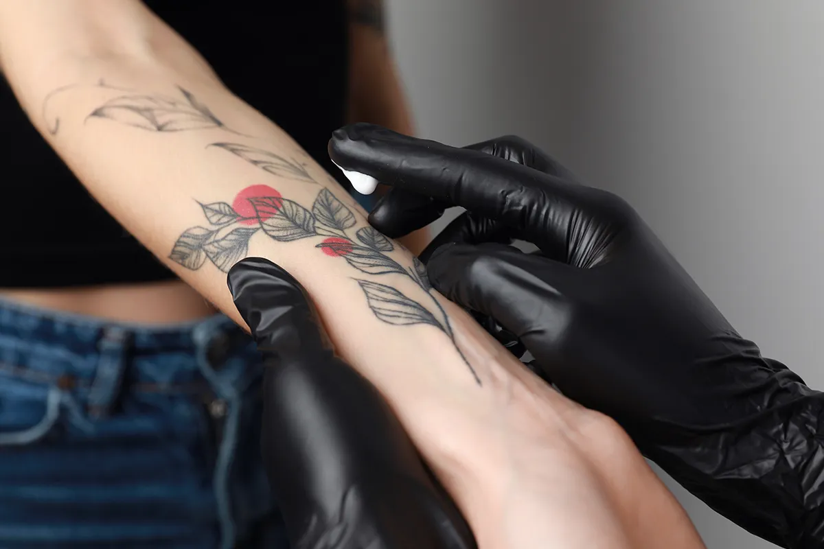 Nahaufnahme von Creme an einer Hand in einem schwarzen Latex-Handschuh über einem Pflanzentattoo auf einem rechten Unterarm